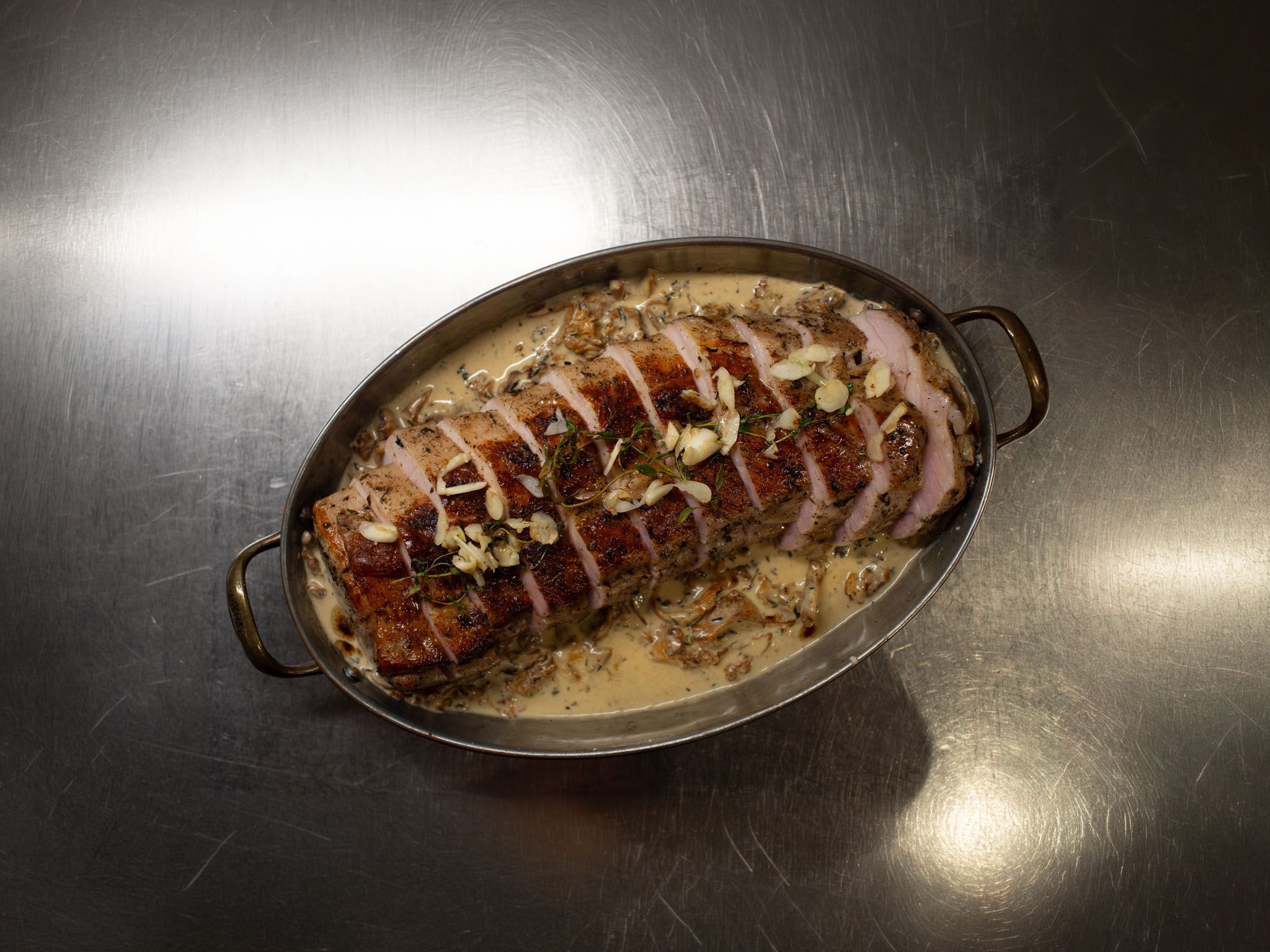 Sliced slow-roasted pork tenderloin in chanterelle cream sauce, topped with garlic and thyme, in an oval pan