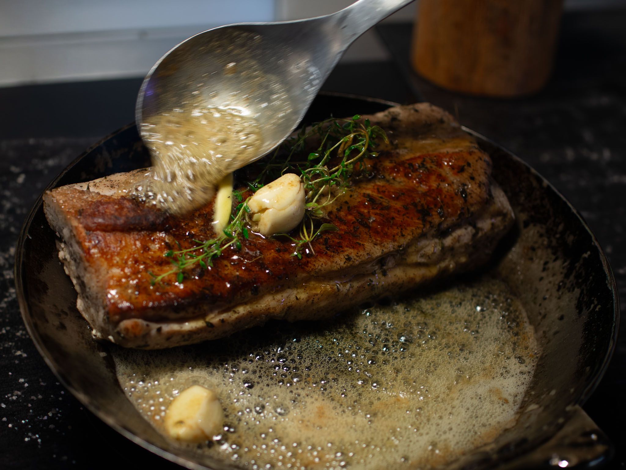 A spoon pouring foaming butter over a seared pork tenderloin in a frying pan, with garlic cloves and sprigs of thyme